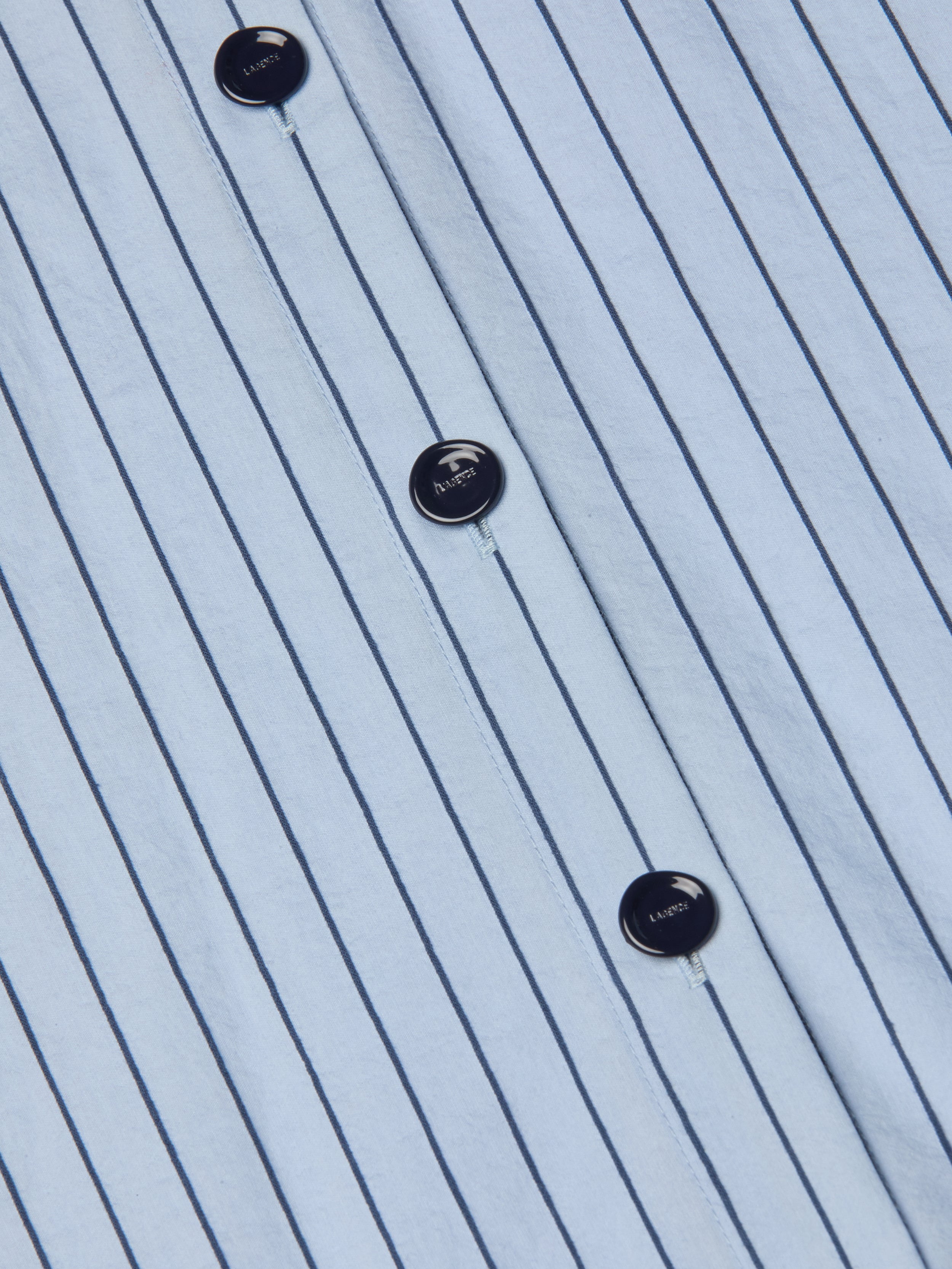 Close-up of the Ralphie Midi Dress, featuring a white fabric with vertical thin black stripes and black buttons, each labeled with LACOSTE and a small crocodile logo. The material appears crisp and neatly pressed.