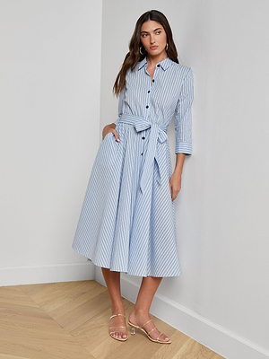 A woman stands indoors against a white wall, wearing the Ralphie Midi Dress—a light blue striped Italian poplin shirt dress with a waist tie—and nude sandals. She has long brown hair and a relaxed posture.