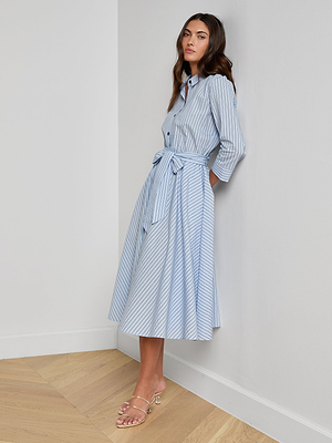 A woman stands against a white wall wearing the Ralphie Midi Dress in light blue and white stripes with a waist tie and clear-strap sandals. The room has light wood flooring.