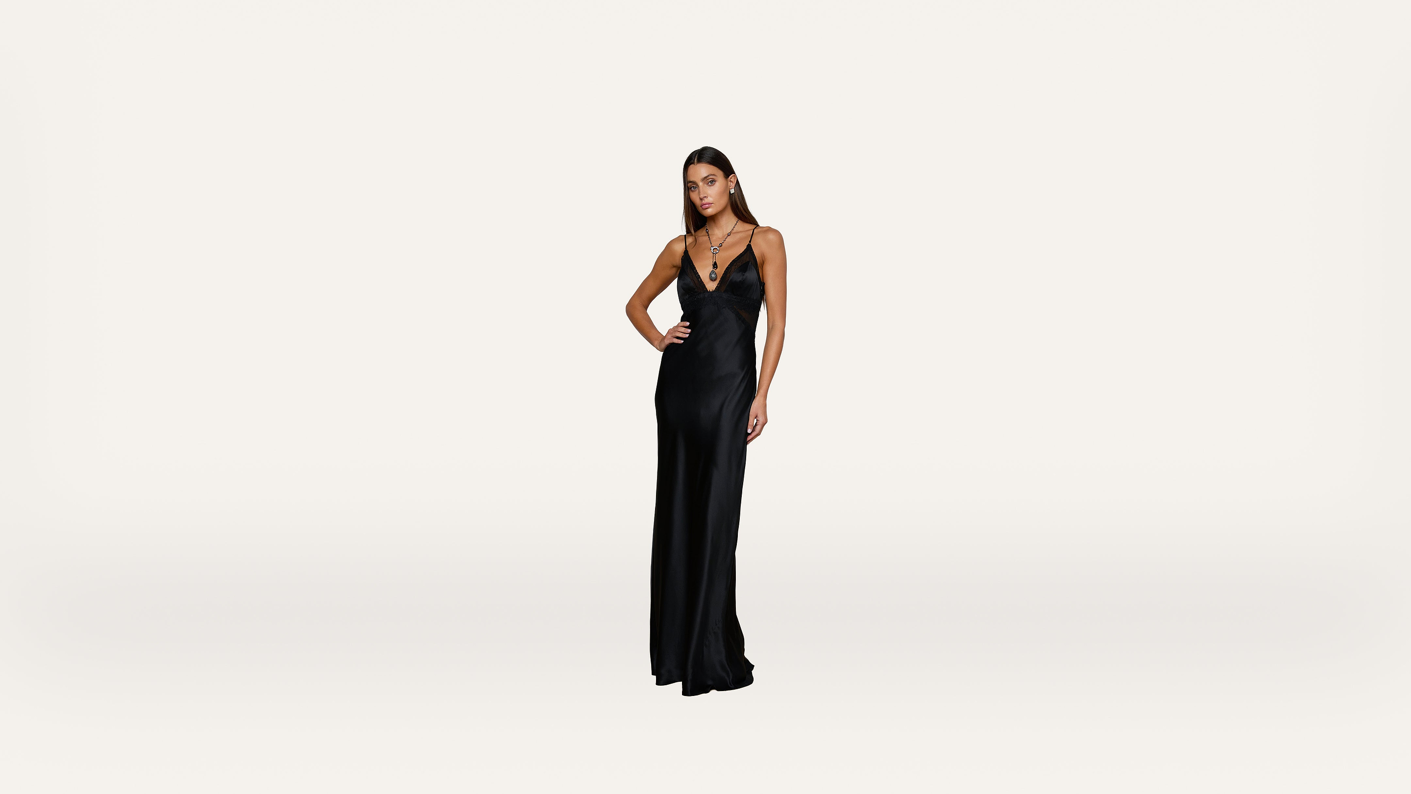 Woman wearing a long, sleeveless black evening gown with a deep V neckline, standing with one hand on her hip against a plain, light background.