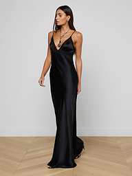 A woman stands on a wooden floor against a plain white wall, wearing the Elsa Silk Gown—a long black slip dress with thin straps—styled with layered necklaces and earrings, looking to her left.