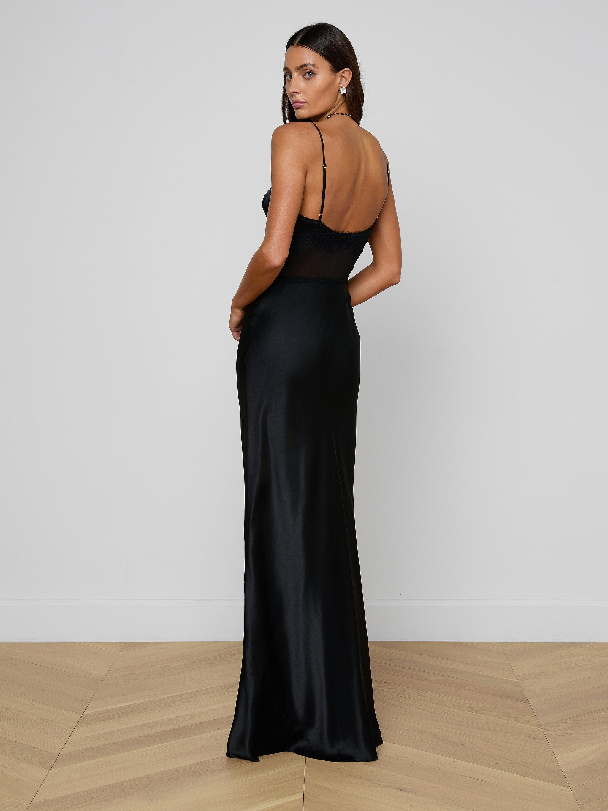 A woman wearing the Elsa Silk Gown, a long, elegant slip dress with thin straps, stands on a wooden floor facing slightly away from the camera and looking over her shoulder against a plain white wall.