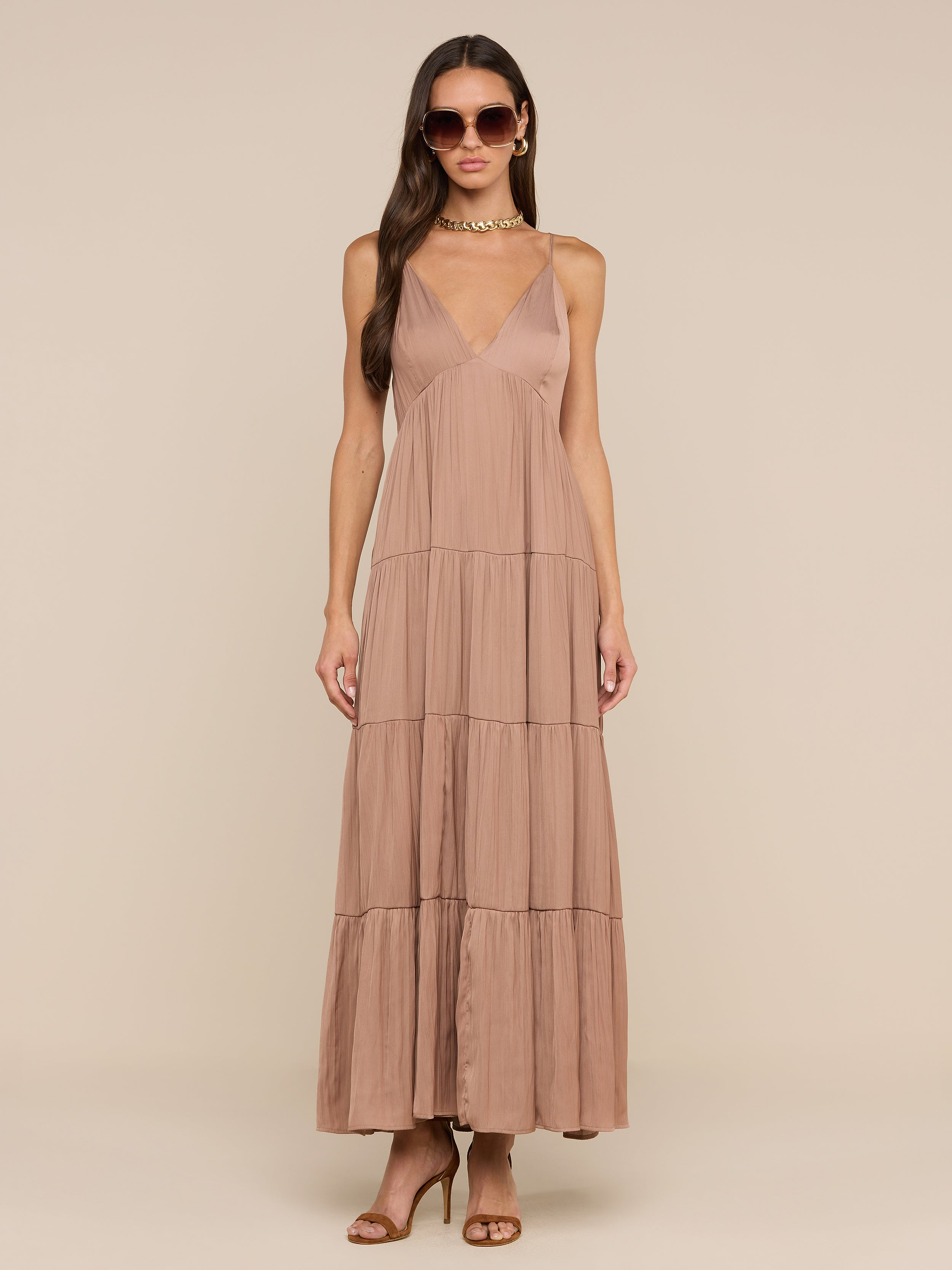 امرأة ترتدي نظارات شمسية وصندل بكعب عالٍ بني اللون وترتدي فستان Lua Tiered Maxi Dress - وهو فستان طويل من الساتان الرمادي الداكن بلا أكمام مع أحزمة قابلة للتعديل وخط رقبة عميق على شكل حرف V وتأثير مجعد وتصميم متدرج - على خلفية باللون البيج.