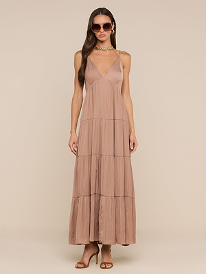 Une femme portant des lunettes de soleil et des sandales à talons marron présente la robe longue Lua Tiered Maxi Dress - une robe longue en satin sans manches de couleur taupe avec des bretelles réglables, une encolure en V profonde, un effet froissé et une silhouette en gradins - sur un fond beige.