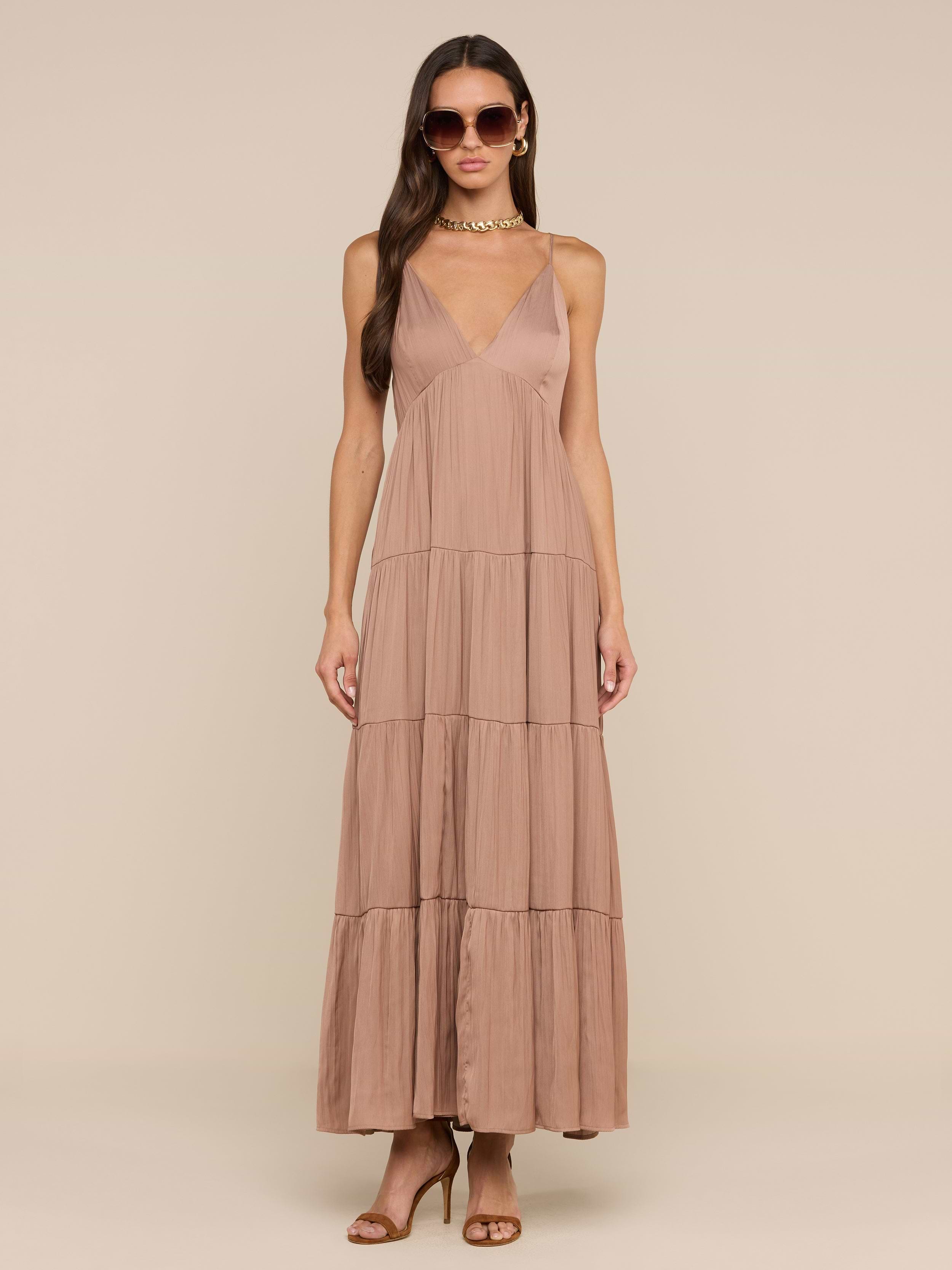 امرأة ترتدي نظارات شمسية وصندل بكعب عالٍ بني اللون وترتدي فستان Lua Tiered Maxi Dress - وهو فستان طويل من الساتان الرمادي الداكن بلا أكمام مع أحزمة قابلة للتعديل وخط رقبة عميق على شكل حرف V وتأثير مجعد وتصميم متدرج - على خلفية باللون البيج.