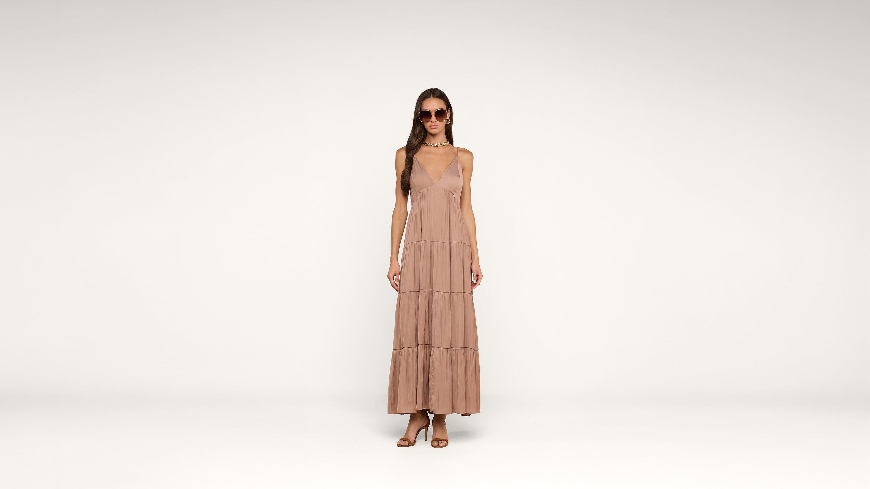 A woman in sunglasses wears a sleeveless, ankle-length blush pink dress and sandals, standing against a plain white background.