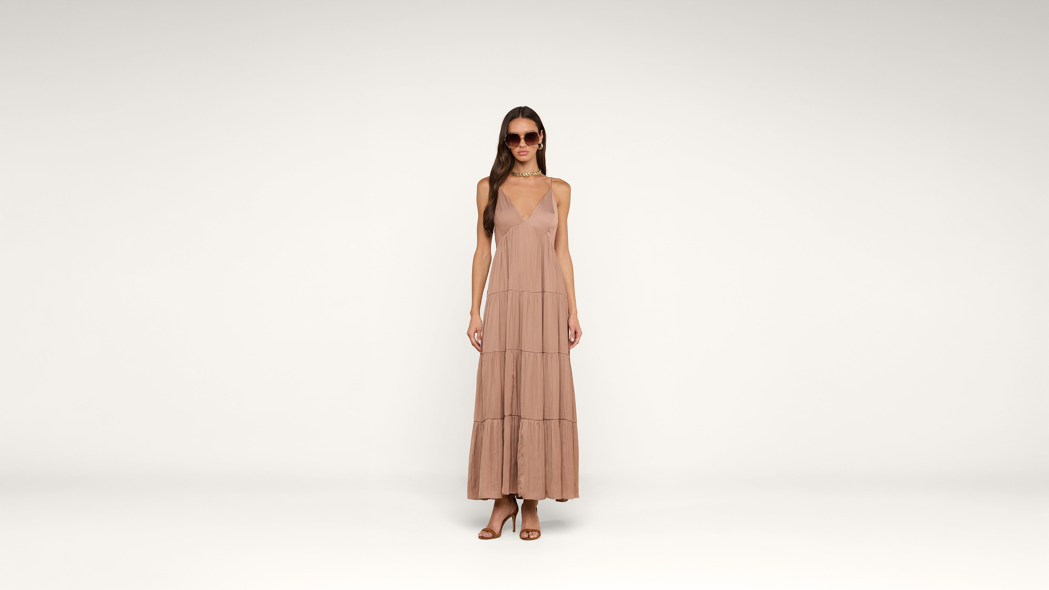 A woman in sunglasses wears a sleeveless, ankle-length blush pink dress and sandals, standing against a plain white background.