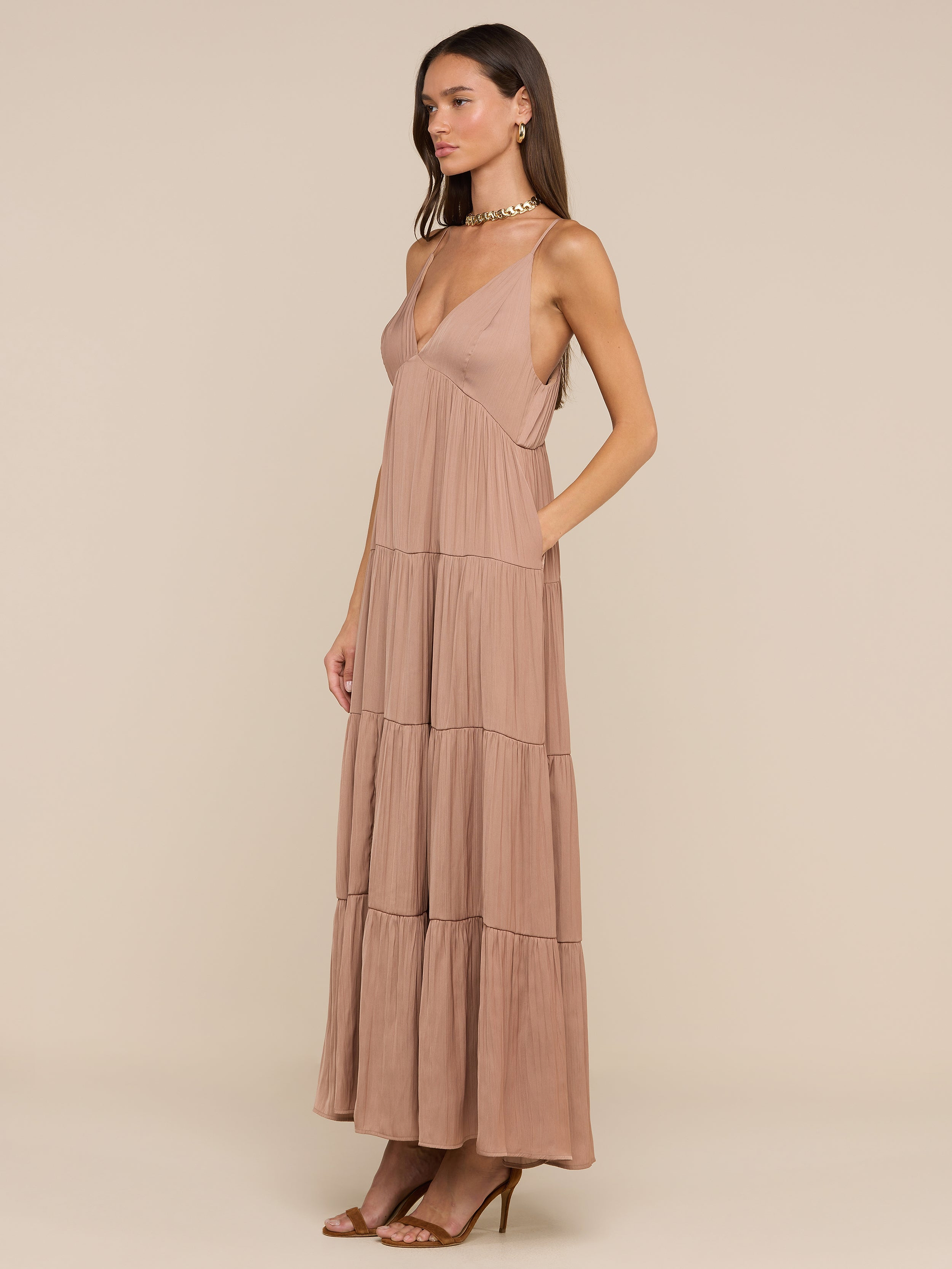 امرأة ترتدي فستان Lua Tiered Maxi Dress - بدون أكمام مع حمالات قابلة للتعديل، وياقة عميقة على شكل V، وياقة على شكل V، وستان مجعد، مع حذاء بكعب عالٍ ذو أحزمة، وتقف على خلفية باللون البيج.