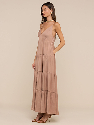 Une femme portant la robe longue Lua Tiered Maxi Dress - sans manches avec des bretelles réglables, un décolleté en V profond et du satin à effet froissé - associée à des talons à lanières, se tient sur un fond beige.