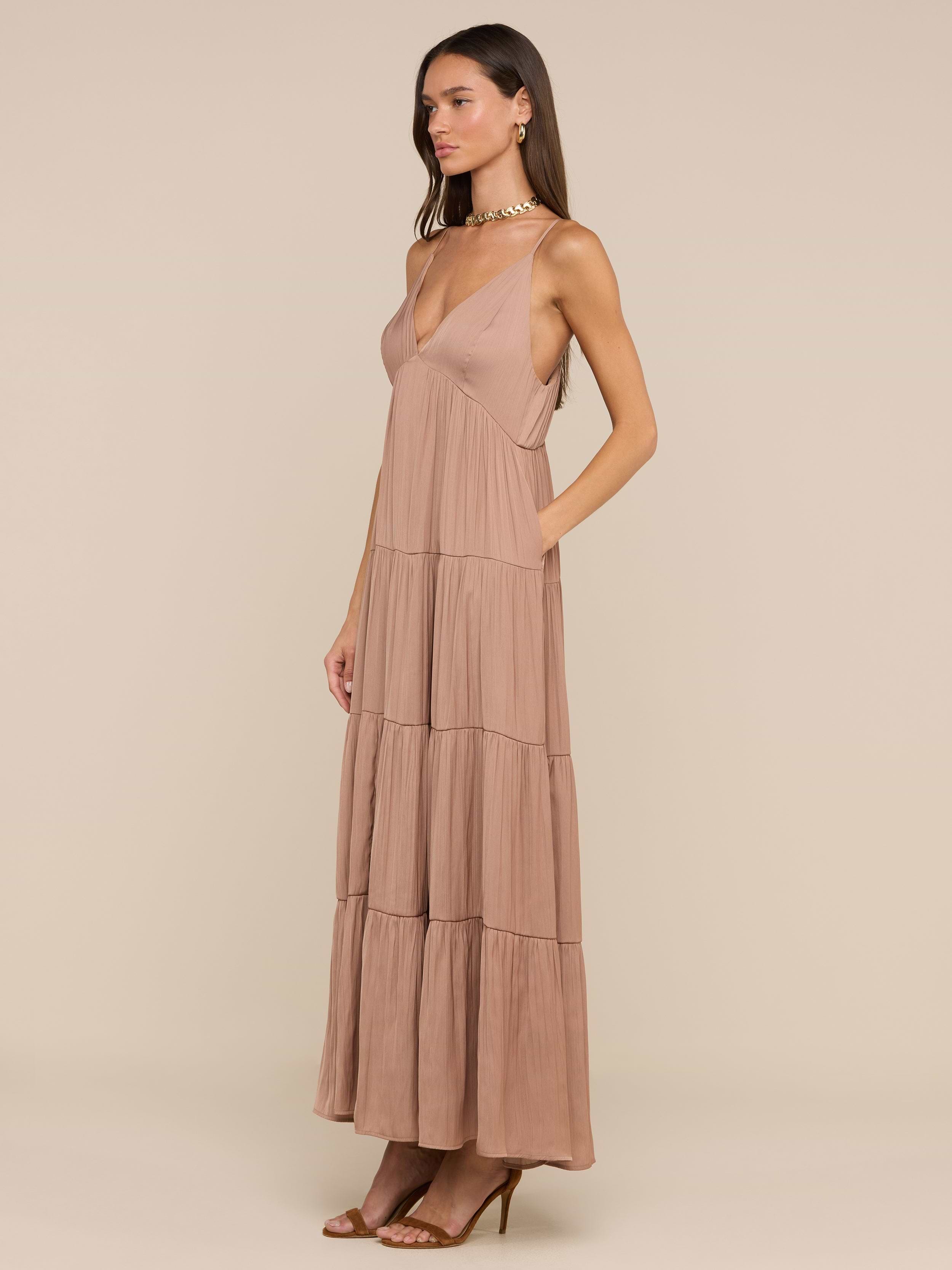 امرأة ترتدي فستان Lua Tiered Maxi Dress - بدون أكمام مع حمالات قابلة للتعديل، وياقة عميقة على شكل V، وياقة على شكل V، وستان مجعد، مع حذاء بكعب عالٍ ذو أحزمة، وتقف على خلفية باللون البيج.