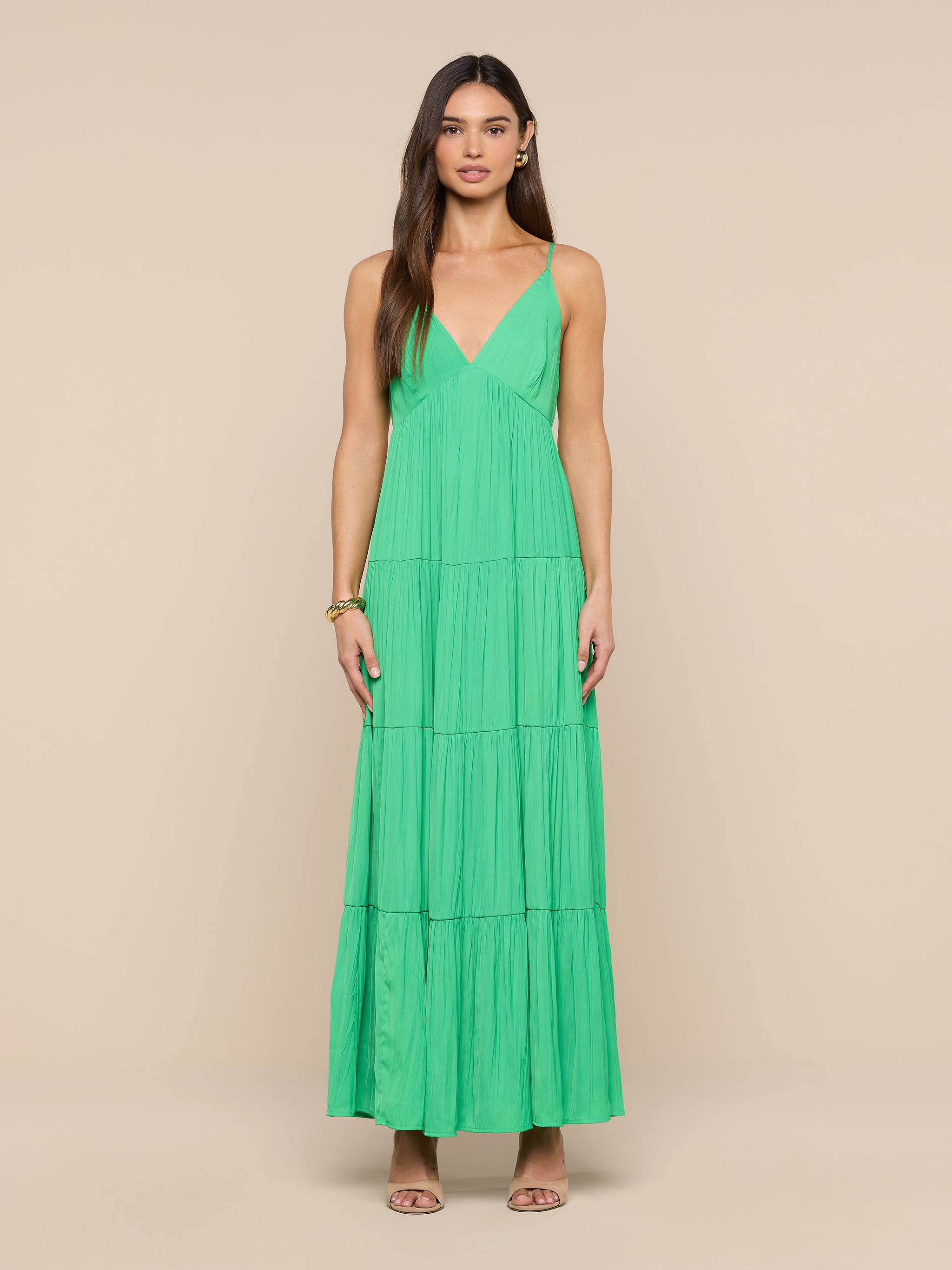 A woman faces forward against a plain white background, wearing the Lua Tiered Maxi Dress—a sleeveless, floor-length bright green maxi dress featuring a V-neckline and adjustable straps. She has long brown hair and accessorizes with sandals and a bracelet.