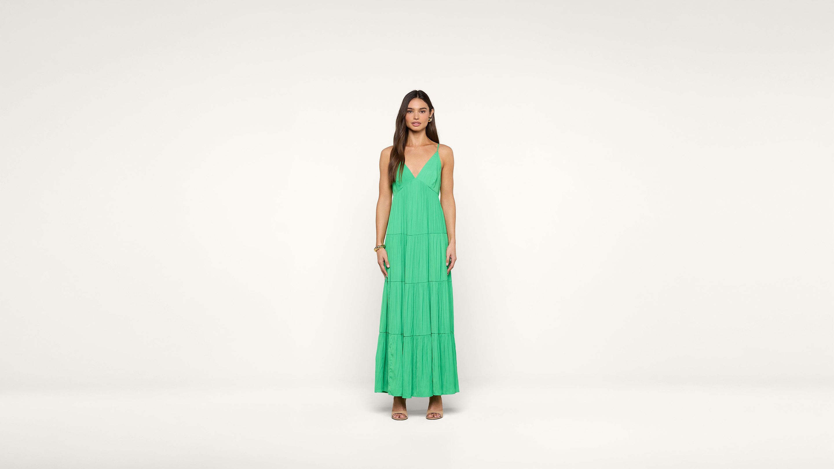 A woman stands against a plain white background wearing a sleeveless green maxi dress with a deep V-neckline and tiered skirt. She has long, straight hair and is wearing sandals.