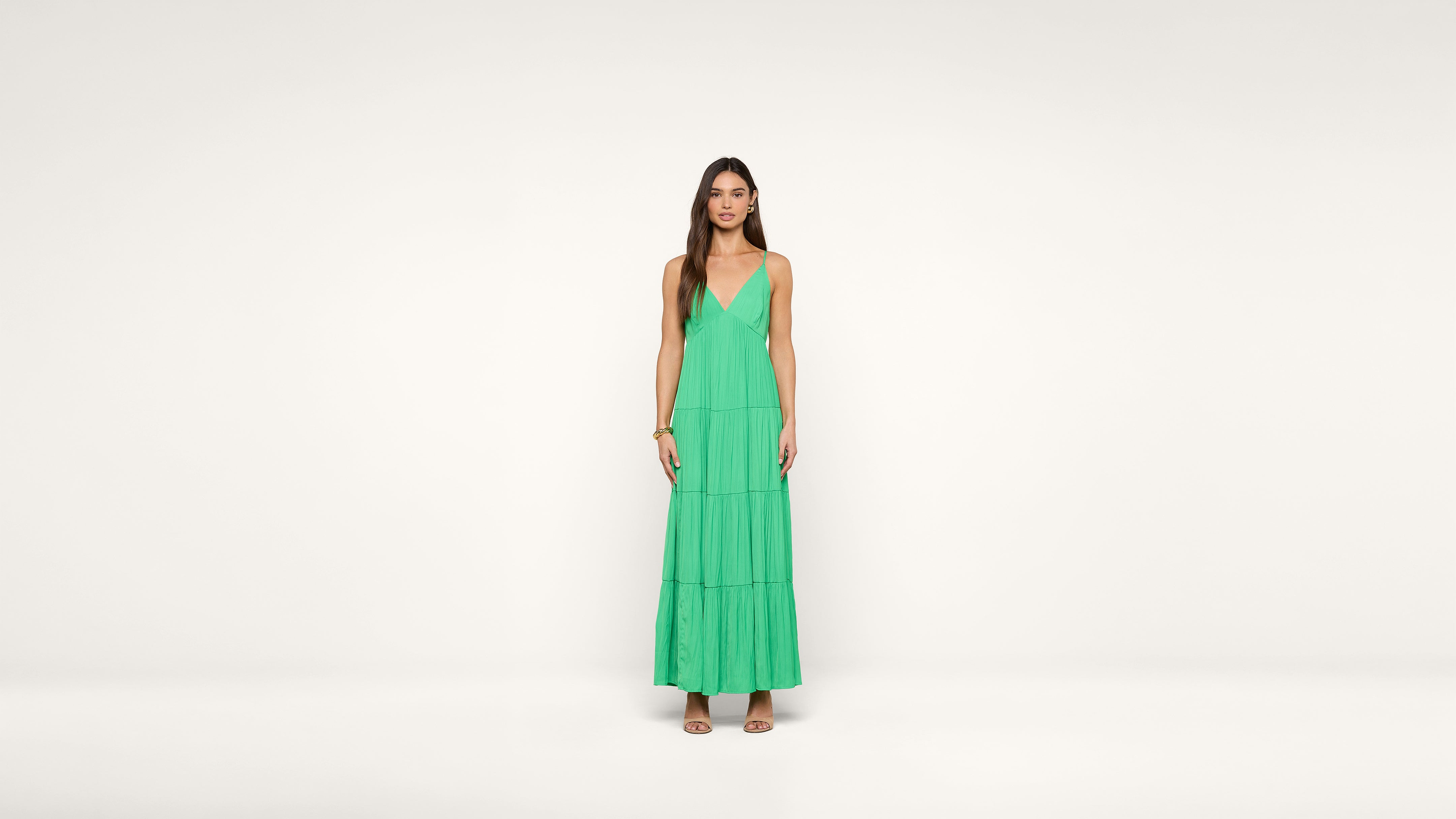 A woman stands against a plain white background wearing a sleeveless green maxi dress with a deep V-neckline and tiered skirt. She has long, straight hair and is wearing sandals.
