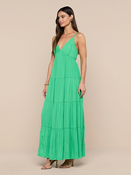 A woman stands against a plain white background, wearing the Lua Tiered Maxi Dress—a sleeveless green maxi dress with adjustable straps and a tiered silhouette. The crinkle-effect satin fabric brings texture and elegance to her look, which she completes with brown heeled sandals.