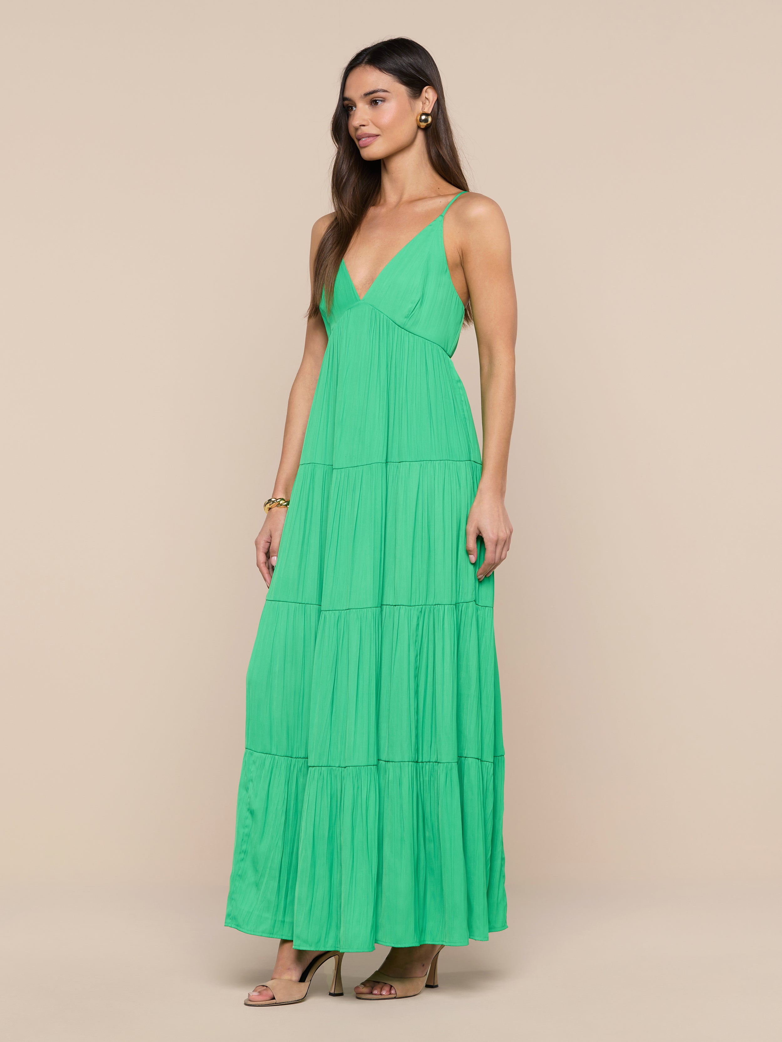 A woman stands against a plain white background, wearing the Lua Tiered Maxi Dress—a sleeveless green maxi dress with adjustable straps and a tiered silhouette. The crinkle-effect satin fabric brings texture and elegance to her look, which she completes with brown heeled sandals.