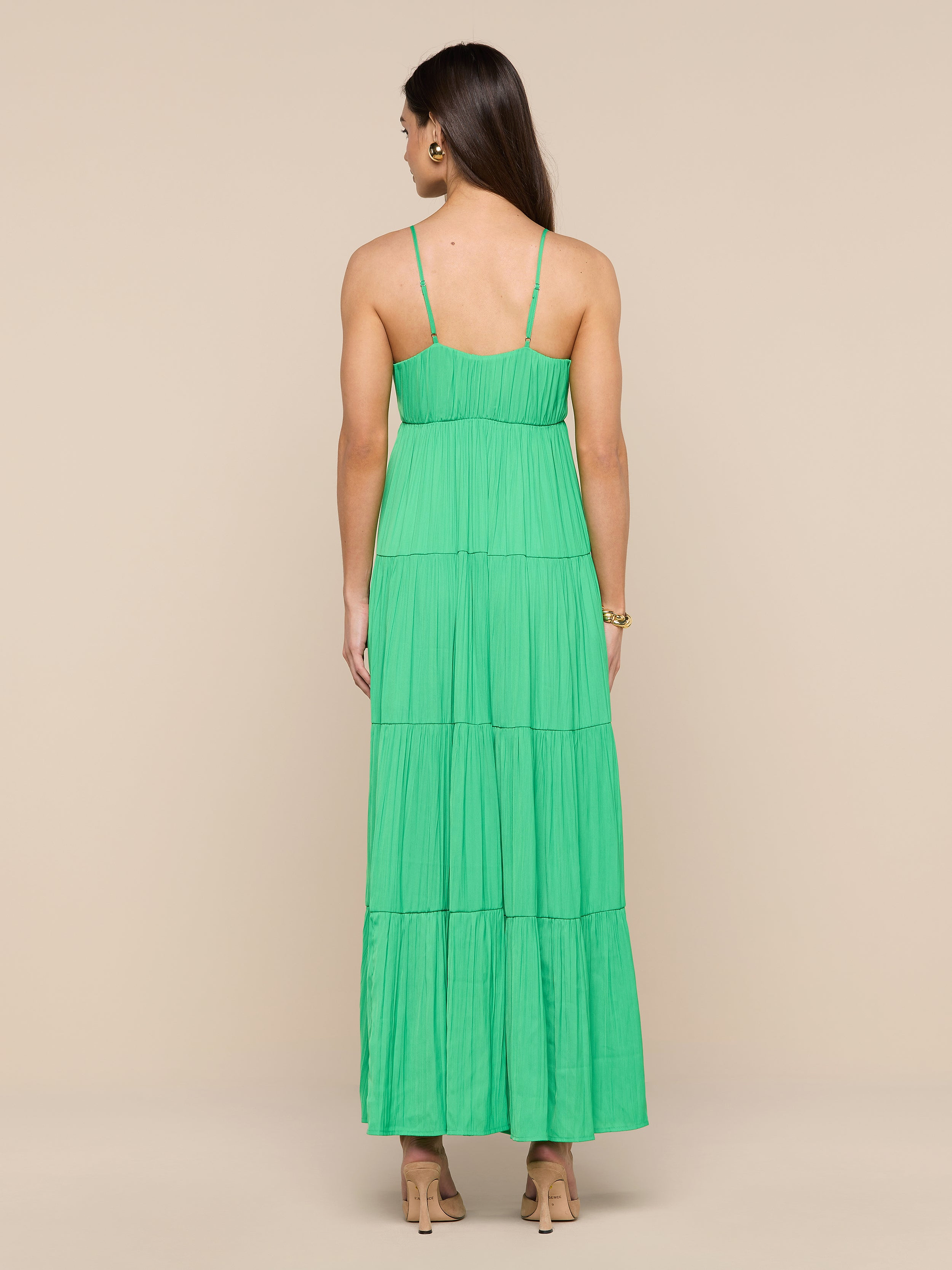 A woman stands facing away, wearing the Lua Tiered Maxi Dress— a sleeveless green maxi dress crafted from crinkle-effect satin with adjustable straps—paired with beige heels, against a plain white background.