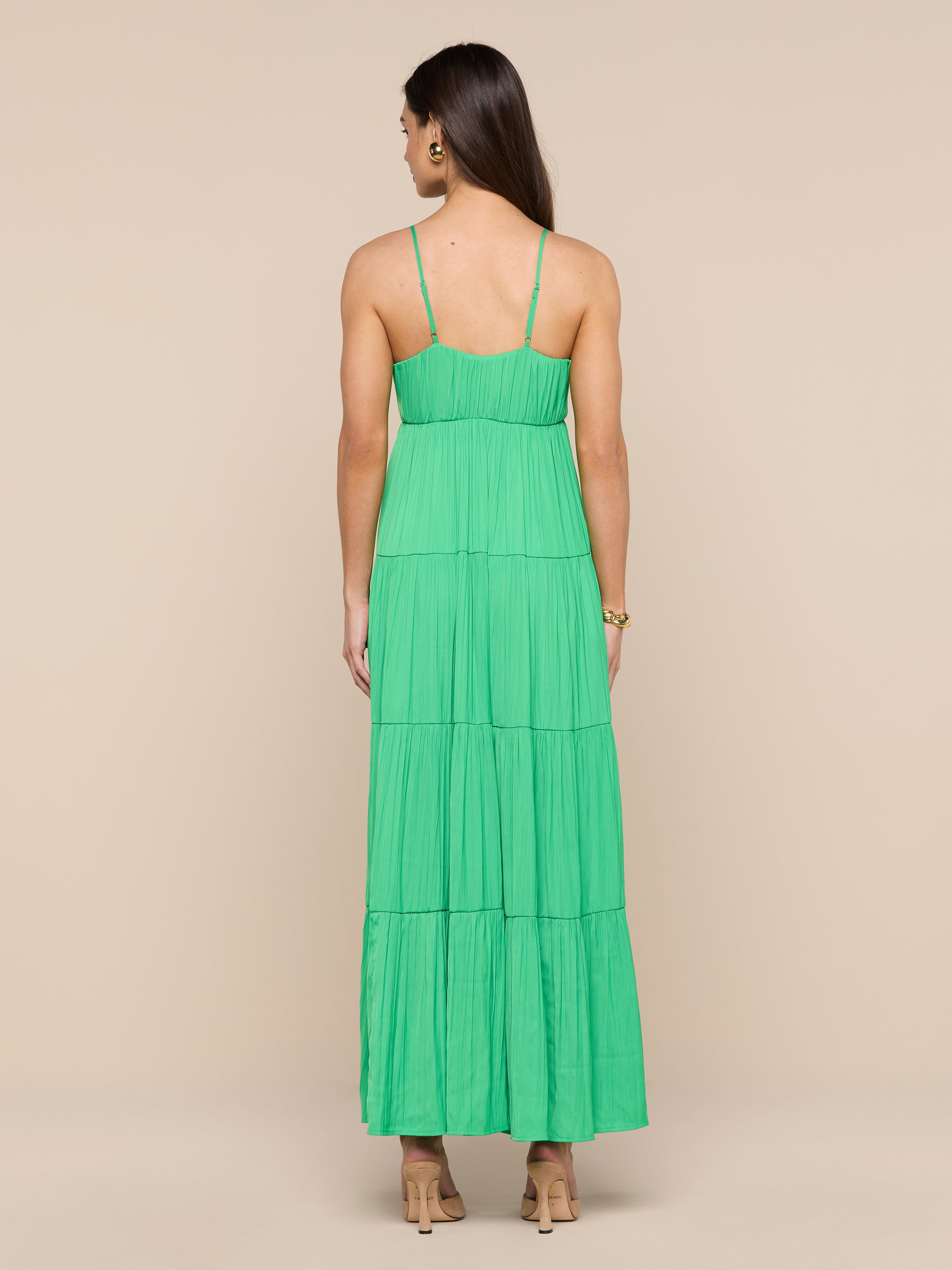 A woman stands facing away, wearing the Lua Tiered Maxi Dress— a sleeveless green maxi dress crafted from crinkle-effect satin with adjustable straps—paired with beige heels, against a plain white background.