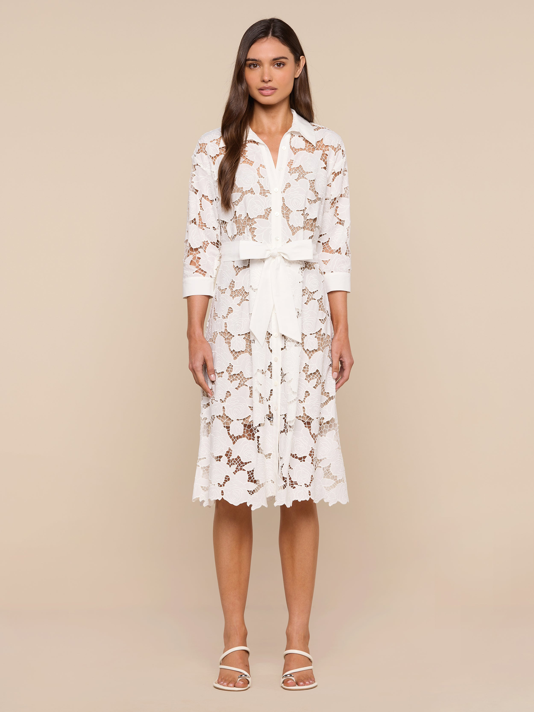 A woman stands against a plain white background, wearing the May Eyelet Shirt Dress—a white cotton midi dress with three-quarter sleeves and a tied waist belt—paired with open-toed white sandals. She has long, straight hair and a neutral expression.