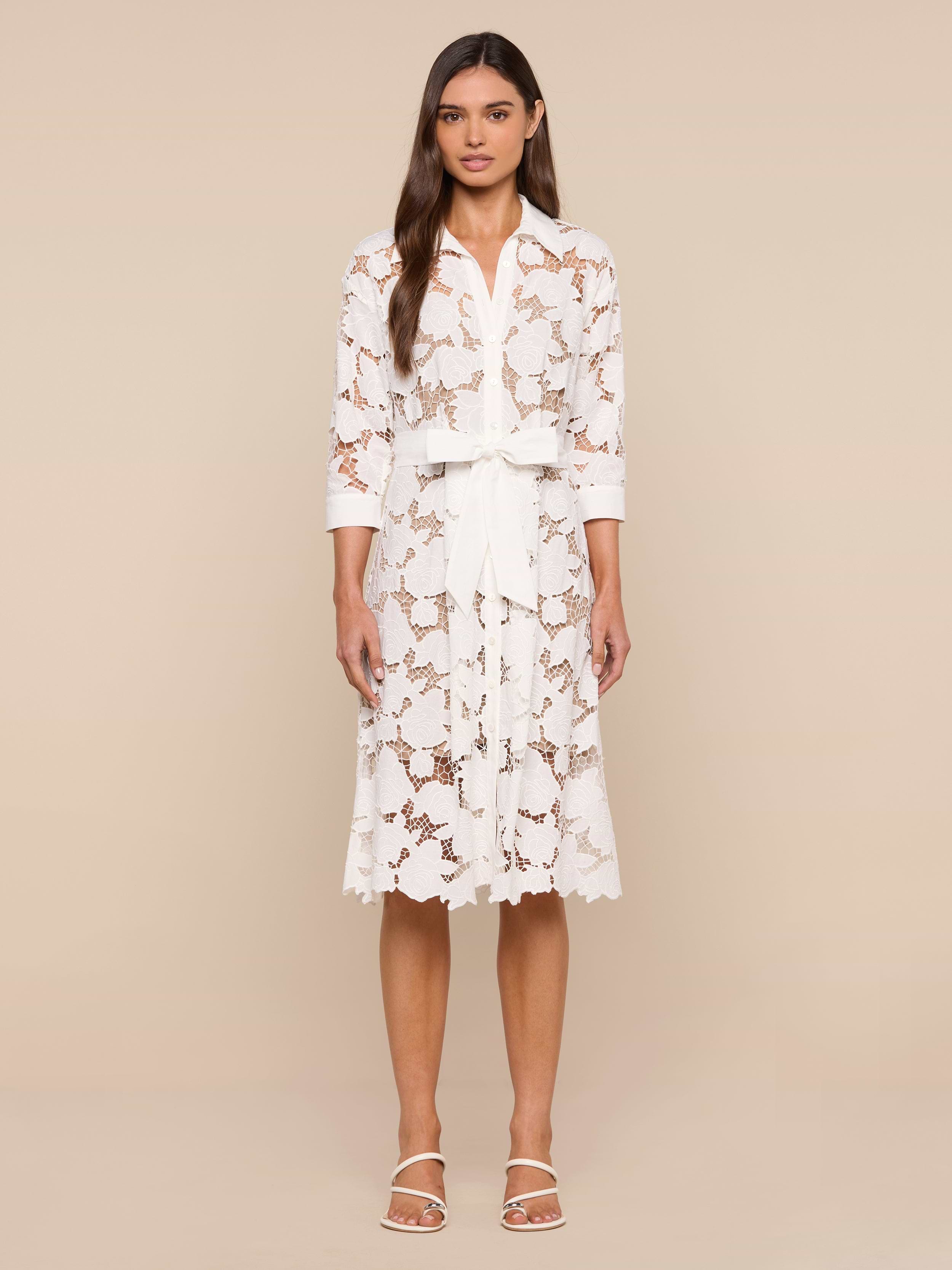 A woman stands against a plain white background, wearing the May Eyelet Shirt Dress—a white cotton midi dress with three-quarter sleeves and a tied waist belt—paired with open-toed white sandals. She has long, straight hair and a neutral expression.