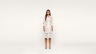 A woman stands facing forward, wearing a white dress with a floral pattern and white sandals, against a plain white background.