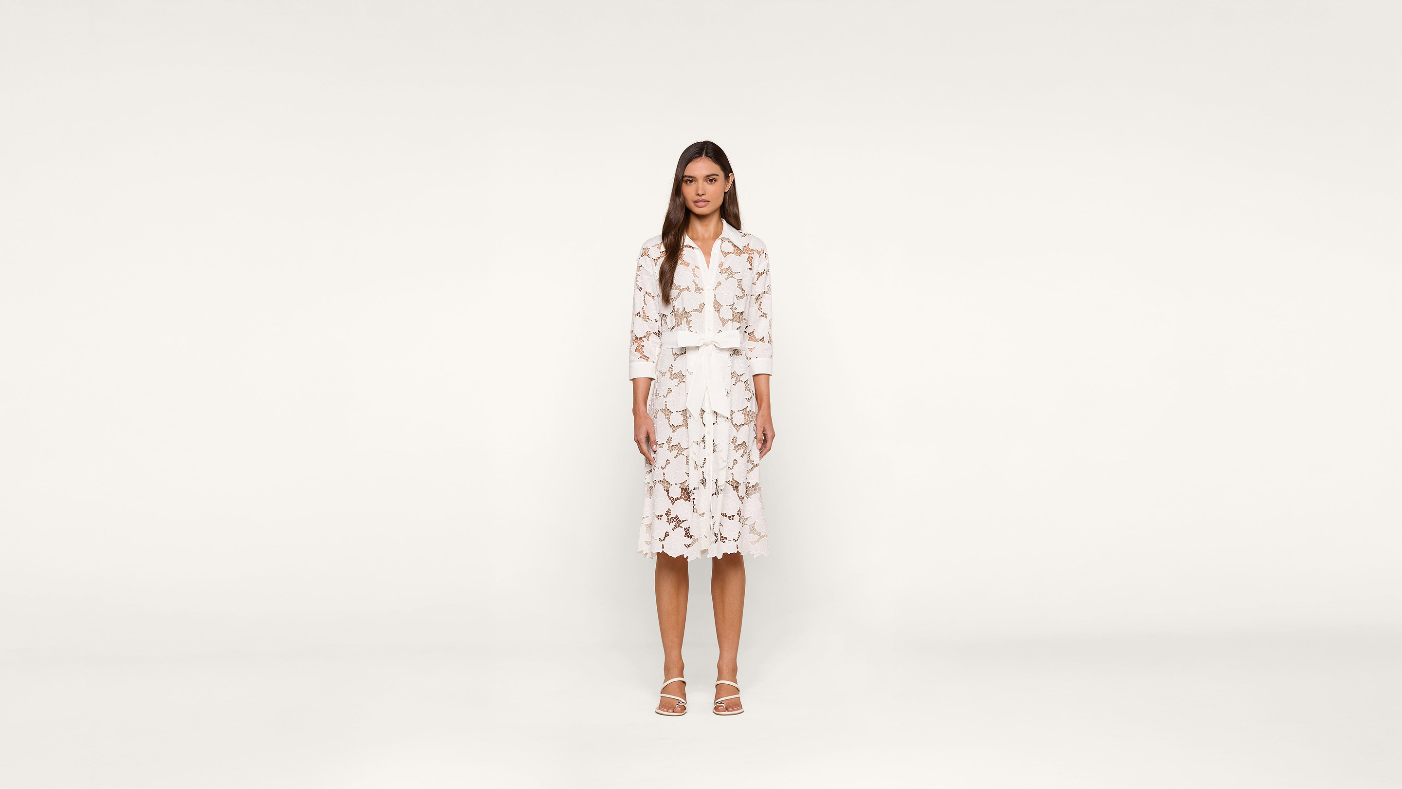 A woman stands facing forward, wearing a white dress with a floral pattern and white sandals, against a plain white background.