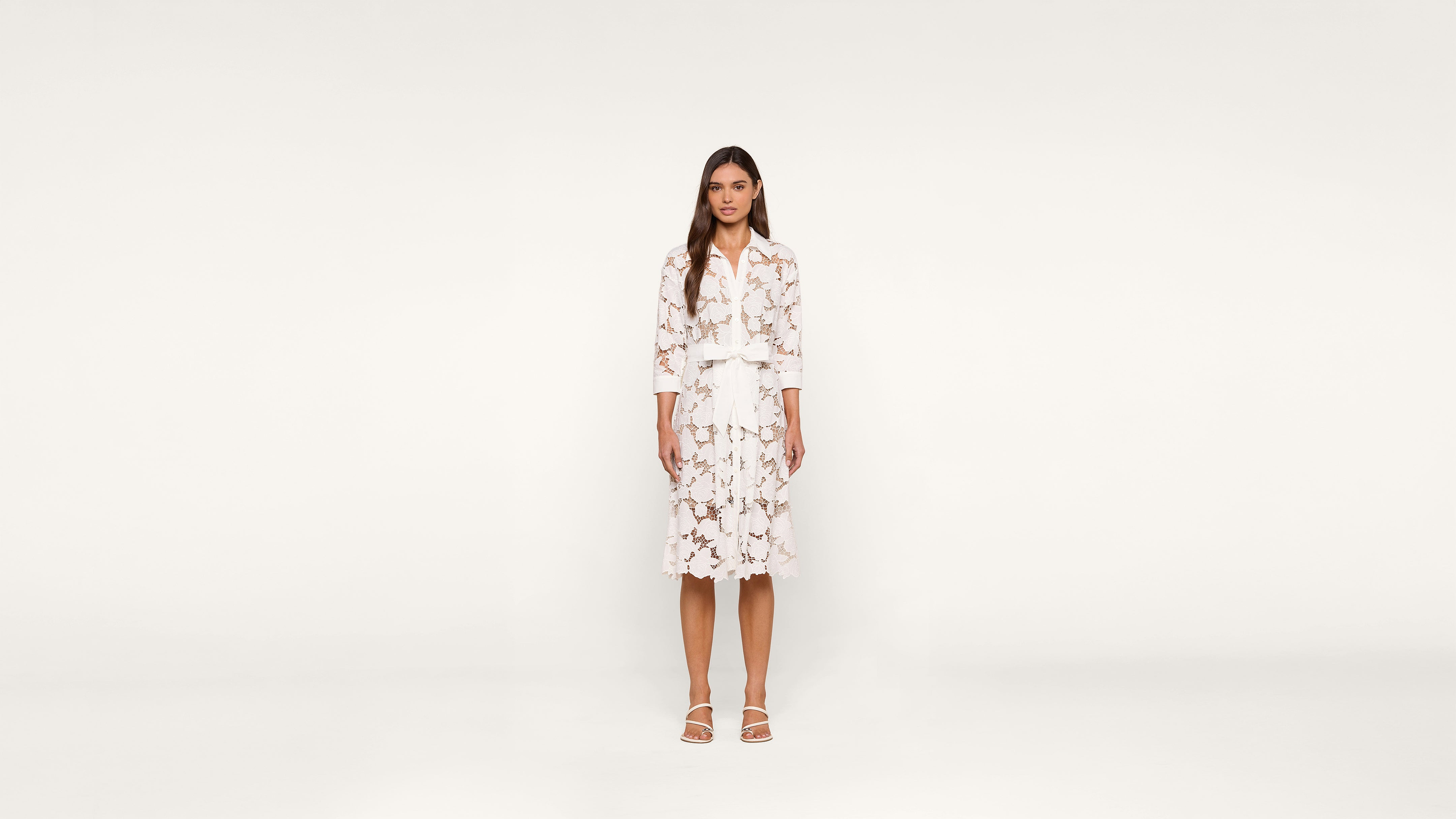 A woman stands facing forward, wearing a white dress with a floral pattern and white sandals, against a plain white background.