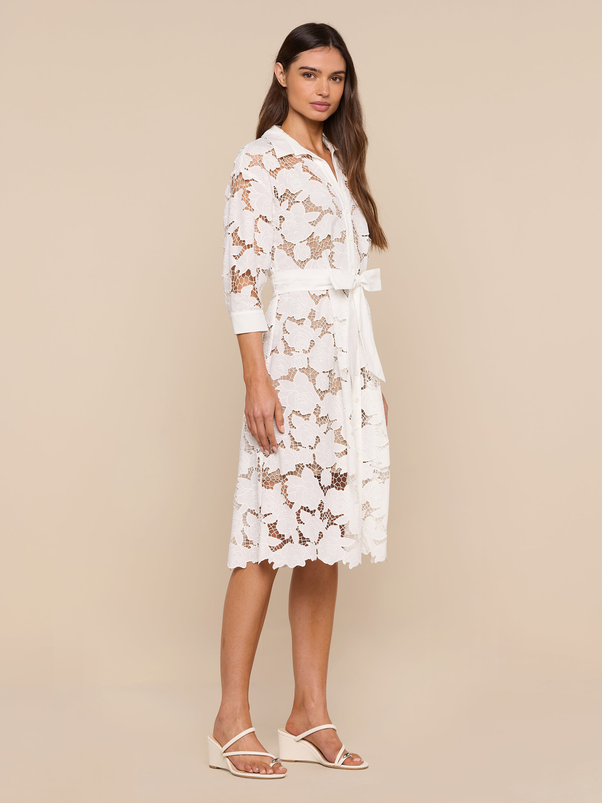 A woman stands against a plain white background wearing the May Eyelet Shirt Dress—a white, knee-length cotton eyelet dress with three-quarter sleeves and a matching belt—paired with white sandals.