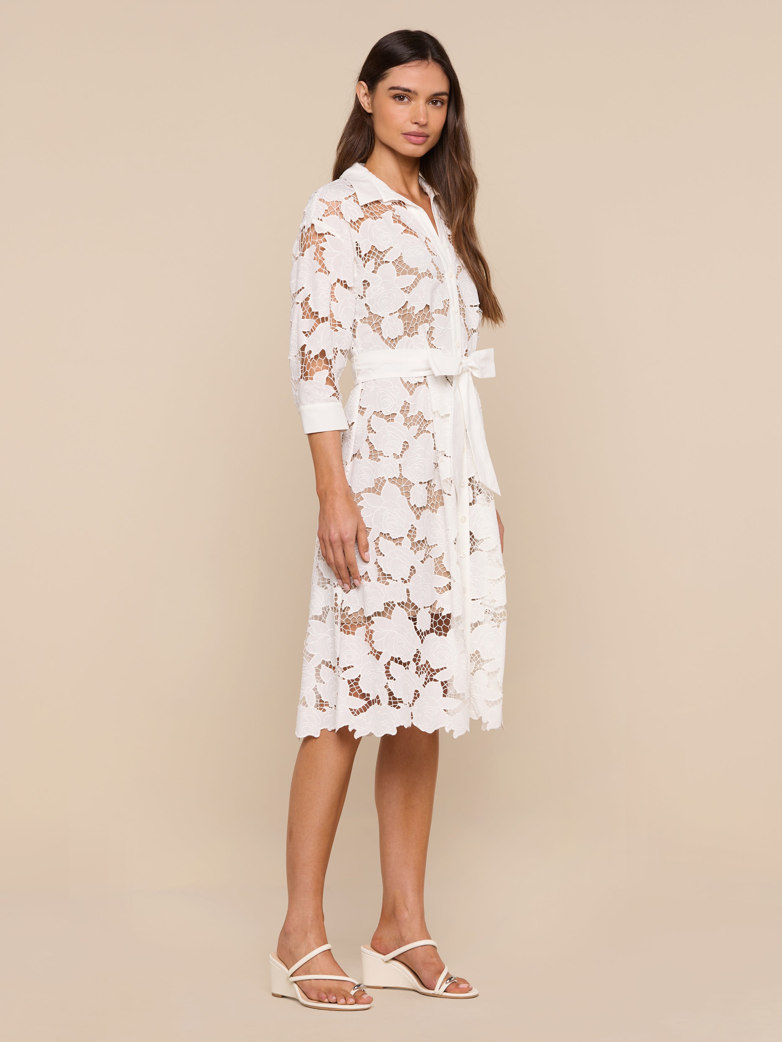 A woman stands against a plain white background wearing the May Eyelet Shirt Dress—a white, knee-length cotton eyelet dress with three-quarter sleeves and a matching belt—paired with white sandals.