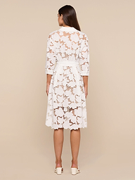 A woman stands facing away, wearing the May Eyelet Shirt Dress—a white, knee-length cotton midi dress with three-quarter sleeves—paired with white heels against a plain, light-colored background.