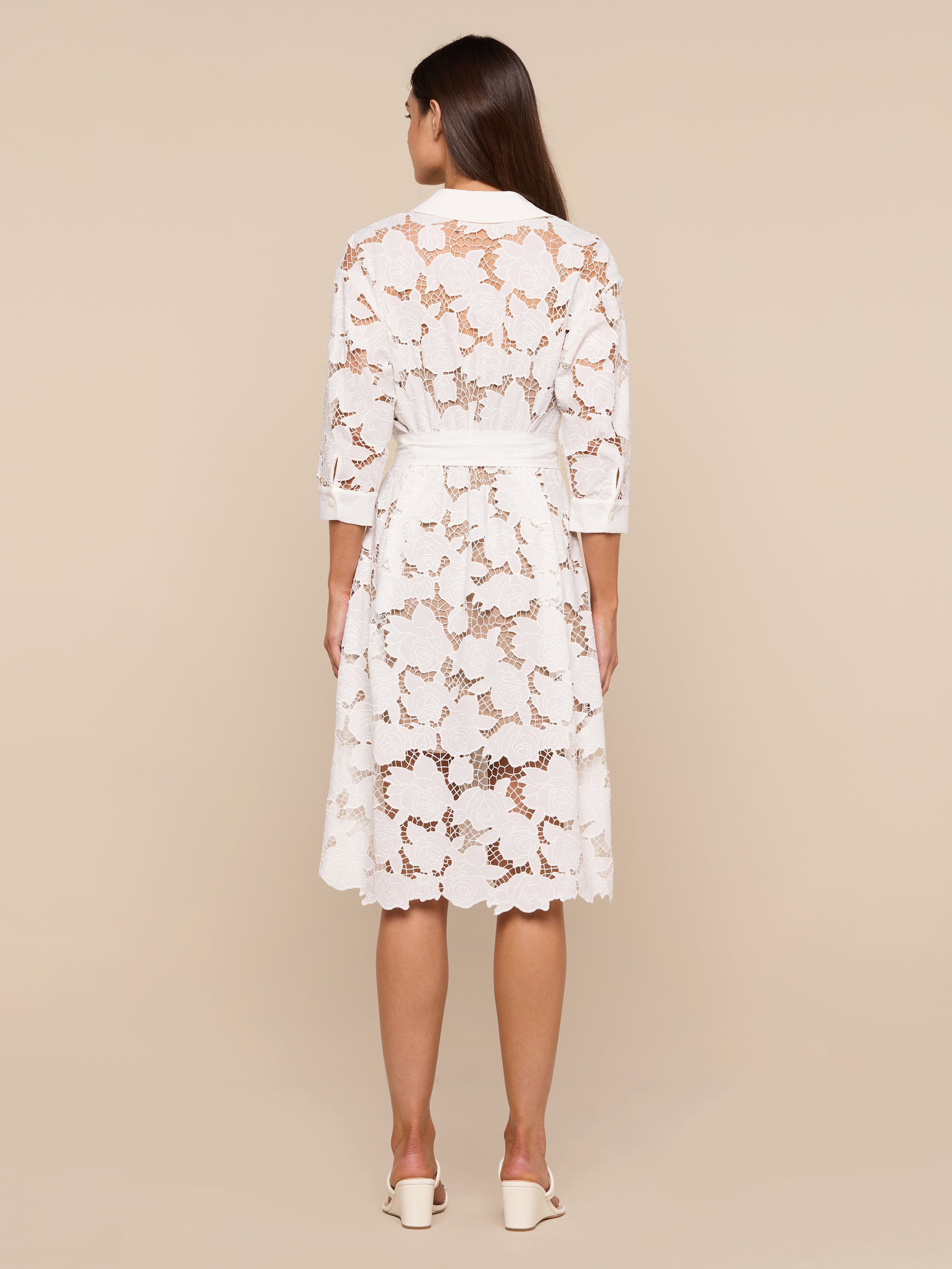 A woman stands facing away, wearing the May Eyelet Shirt Dress—a white, knee-length cotton midi dress with three-quarter sleeves—paired with white heels against a plain, light-colored background.