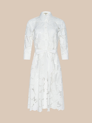 The May Eyelet Shirt Dress is a white cotton midi with long sleeves, button-down front, collar, matching fabric belt at the waist, floral eyelet pattern, and knee length. Displayed on a plain beige background.