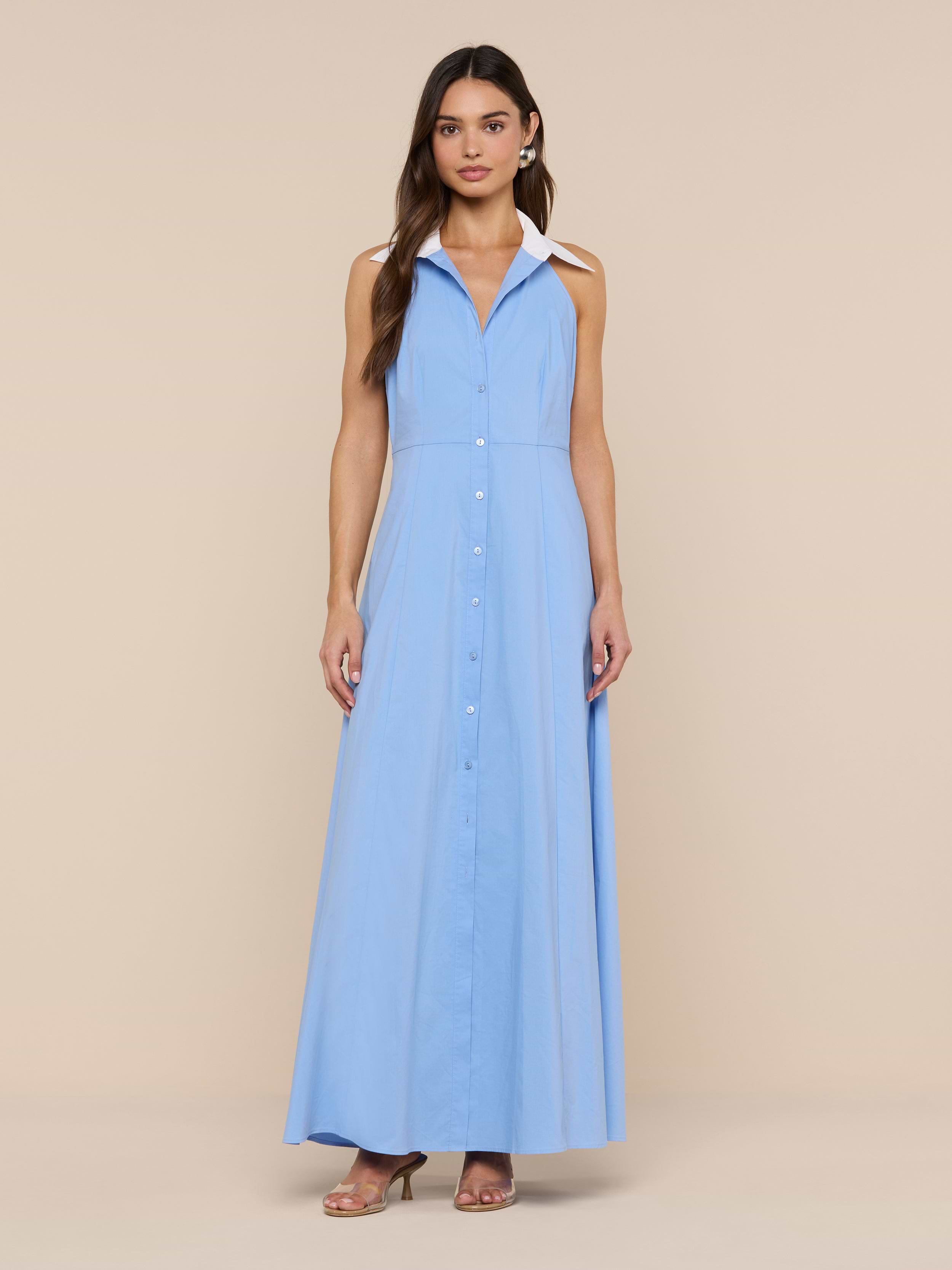 A woman wears the sleeveless, light blue Maurie Poplin Maxi Dress with a collar, crafted from an organic cotton-blend fabric, standing against a plain white background and paired with brown heeled sandals.
