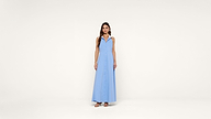 A woman stands facing forward, wearing a sleeveless, light blue, button-down, floor-length dress with a collar and open neckline, paired with sandals, against a plain white background.