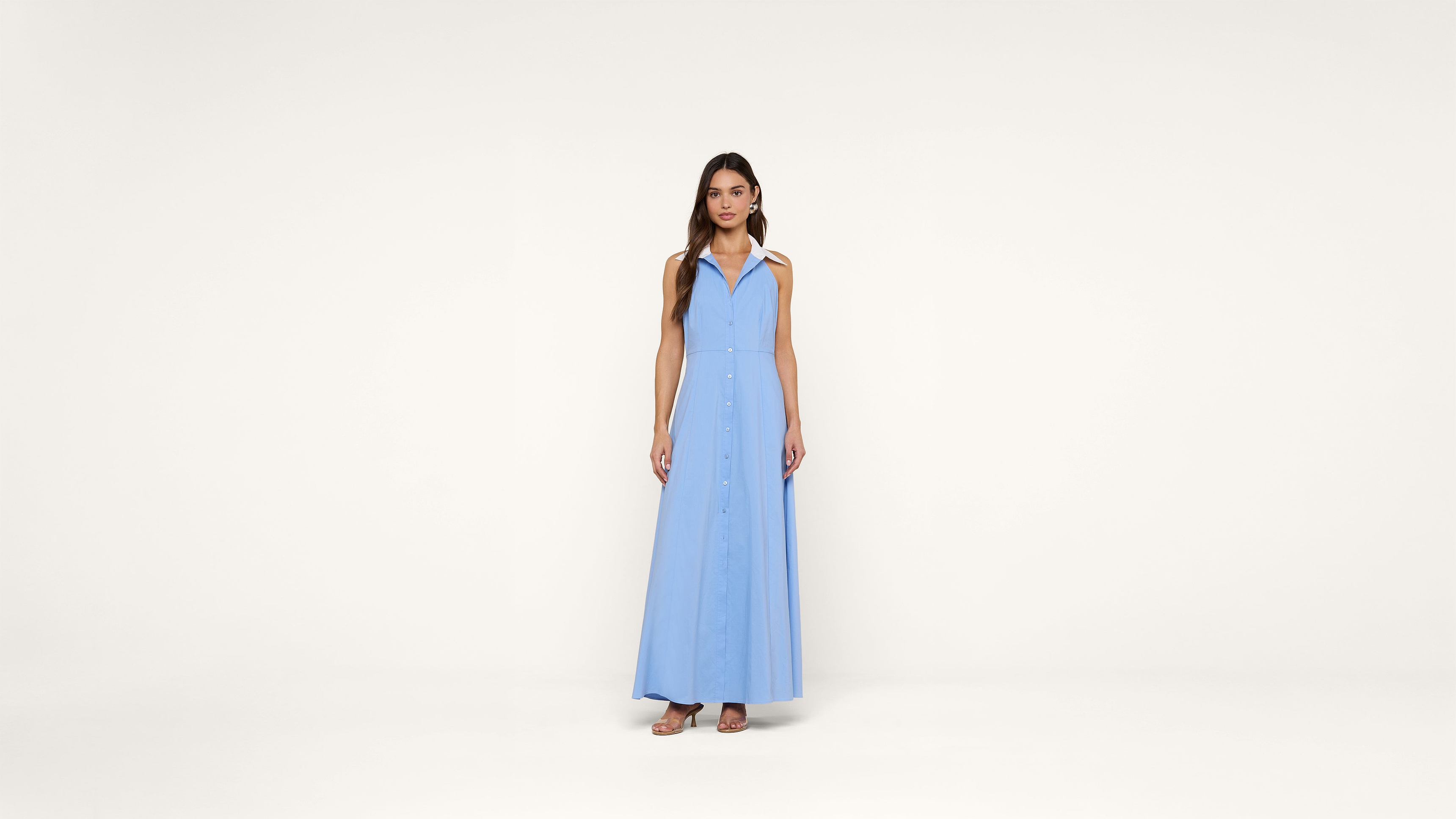 A woman stands facing forward, wearing a sleeveless, light blue, button-down, floor-length dress with a collar and open neckline, paired with sandals, against a plain white background.