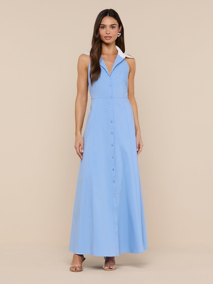 A woman with long brown hair stands against a beige background, wearing the Maurie Poplin Maxi Dress—a light blue organic cotton maxi with a white collar—paired with minimal jewelry and clear-heeled sandals.