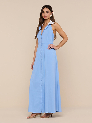 A woman with long brown hair stands against a beige background, wearing the Maurie Poplin Maxi Dress—a sleeveless, light blue organic cotton poplin maxi dress with a collar and clear-heeled sandals.