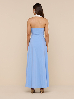 A woman with long brown hair, in beige high-heeled shoes, stands facing away against a beige background wearing the Maurie Poplin Maxi Dress—a light blue organic cotton poplin maxi dress with a white halter neck strap.