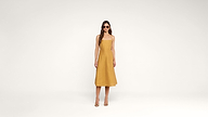 A woman wearing a mustard yellow sleeveless dress and sunglasses stands against a plain white background, looking forward with her arms relaxed at her sides.