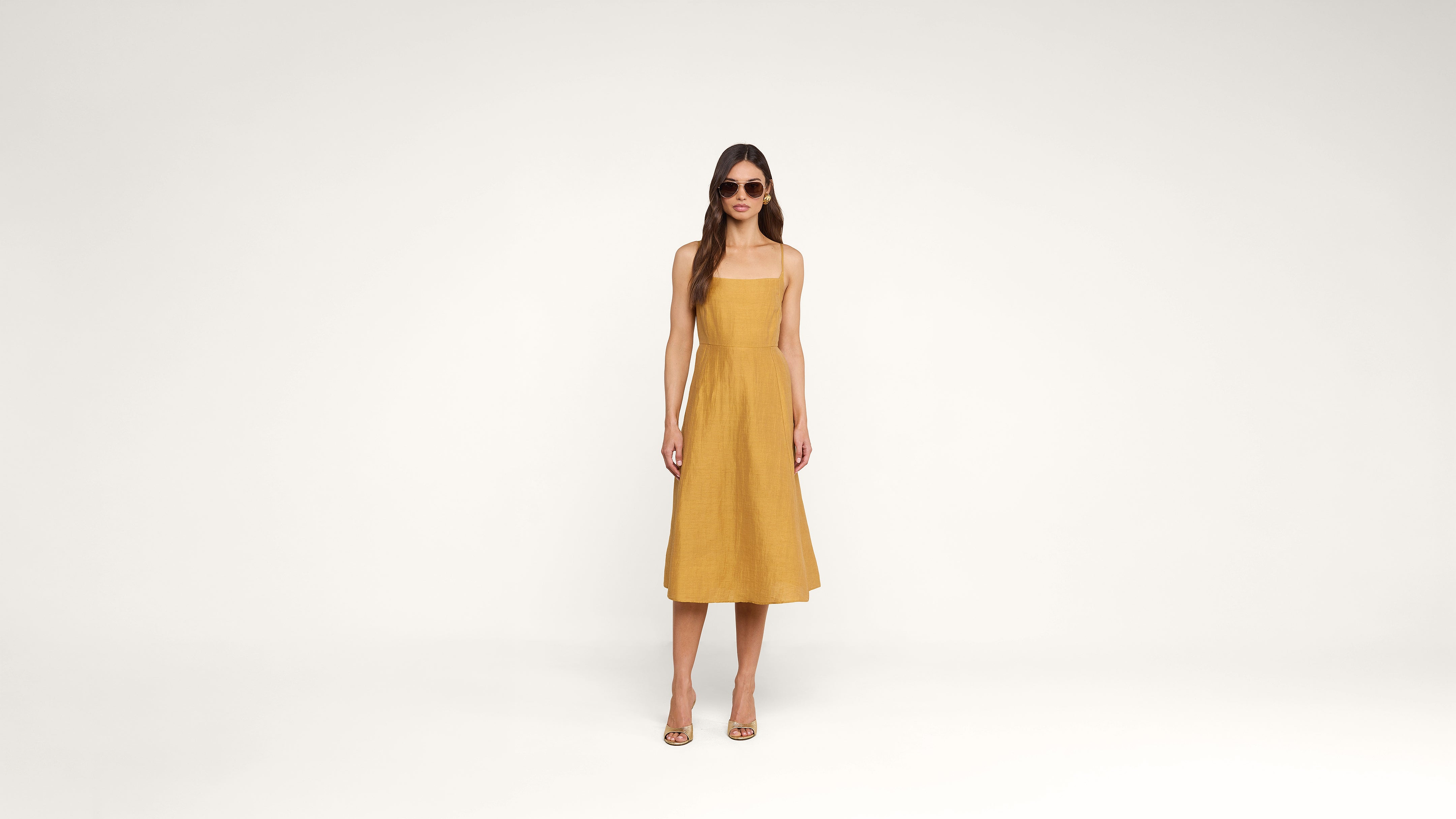 A woman wearing a mustard yellow sleeveless dress and sunglasses stands against a plain white background, looking forward with her arms relaxed at her sides.