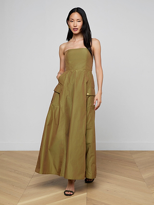 A woman with long dark hair wears the Nessa Strapless Gown in olive green, featuring large front pockets, paired with black open-toe sandals. She stands on a light wood floor against a plain white wall.