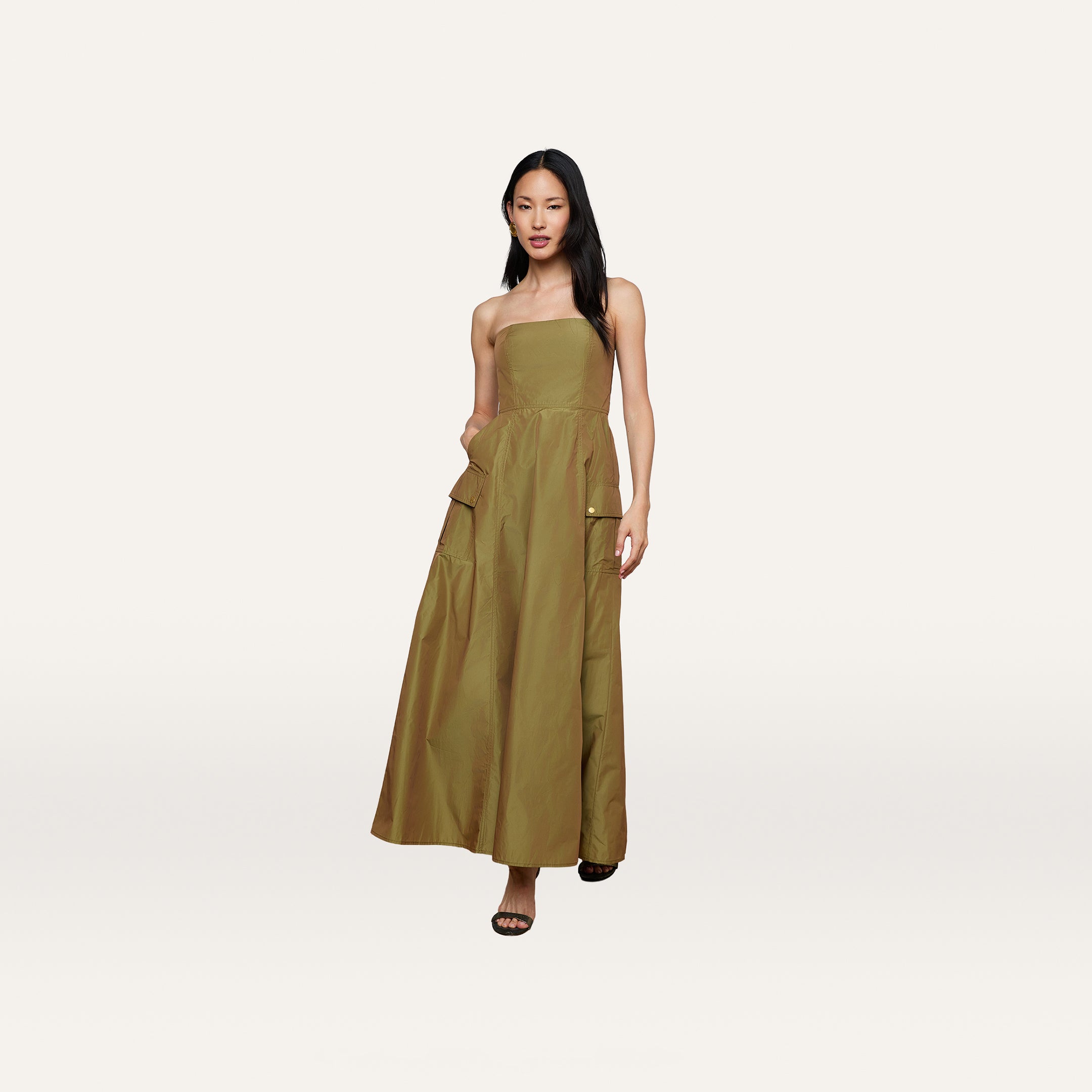 A woman wearing the Nessa Strapless Gown in olive green, featuring a floor-length silhouette and large pockets, stands against a plain white background.