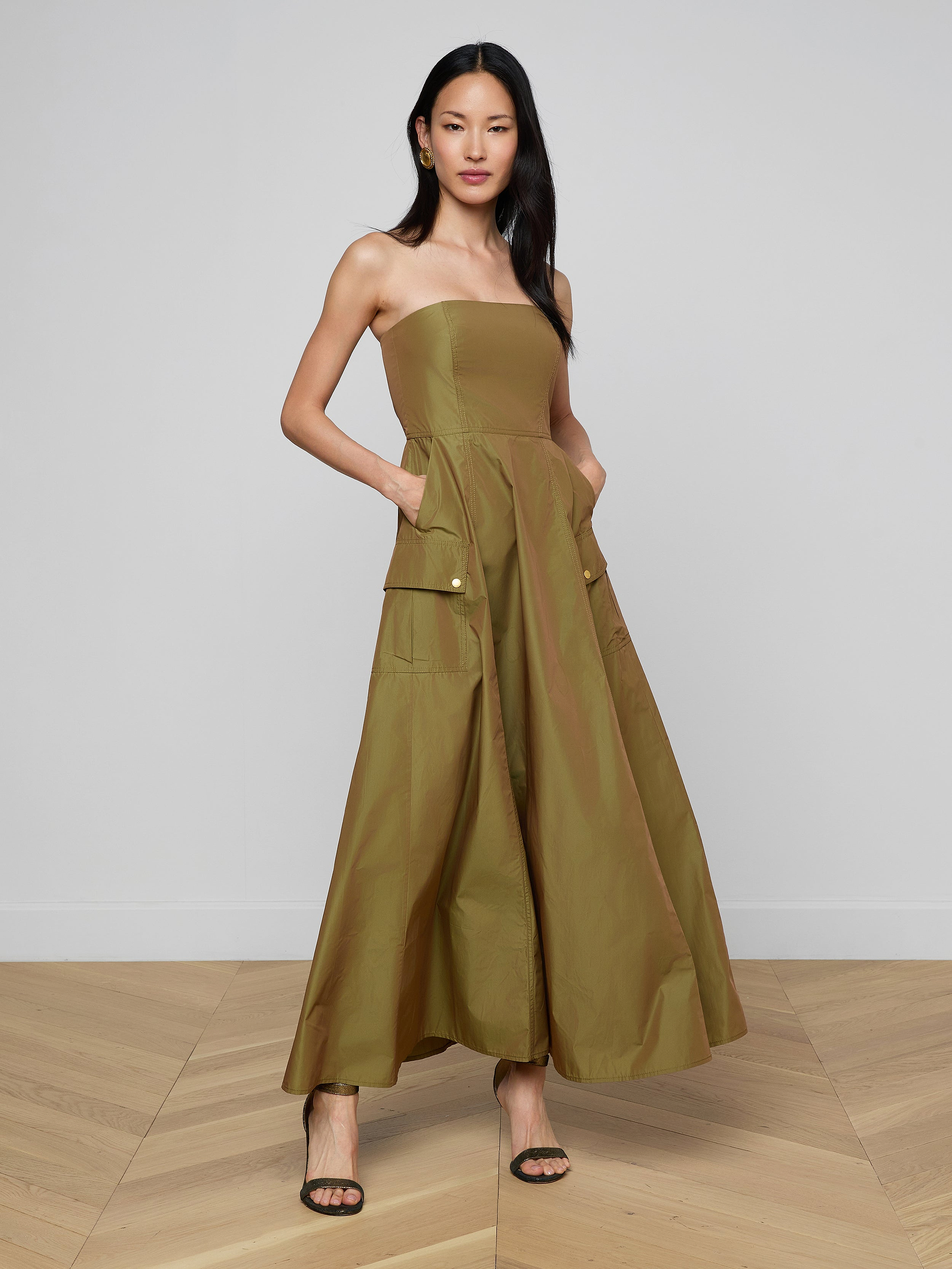 A woman models the Nessa Strapless Gown—an olive green, wide-leg jumpsuit with large pockets and contemporary, floor-length gown-inspired lines—standing with her hands in the pockets and wearing black high-heeled sandals against a plain white background.