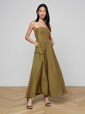 A woman models the Nessa Strapless Gown in olive green—a floor-length dress with large front pockets—paired with black high-heeled sandals. She stands on light wood flooring against a plain white wall, her straight dark hair down.