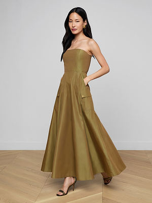 A woman with long dark hair and heeled sandals poses on a wooden floor, wearing the Nessa Strapless Gown—an olive green, floor-length dress with pockets—one hand in her pocket and the other by her side.