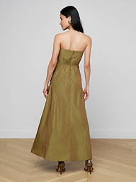 A woman stands facing away, wearing the Nessa Strapless Gown—an olive-green, floor-length dress featuring a fitted bodice and a flared skirt. She has long dark hair, visible earrings, and brown heels, set against a plain white background.