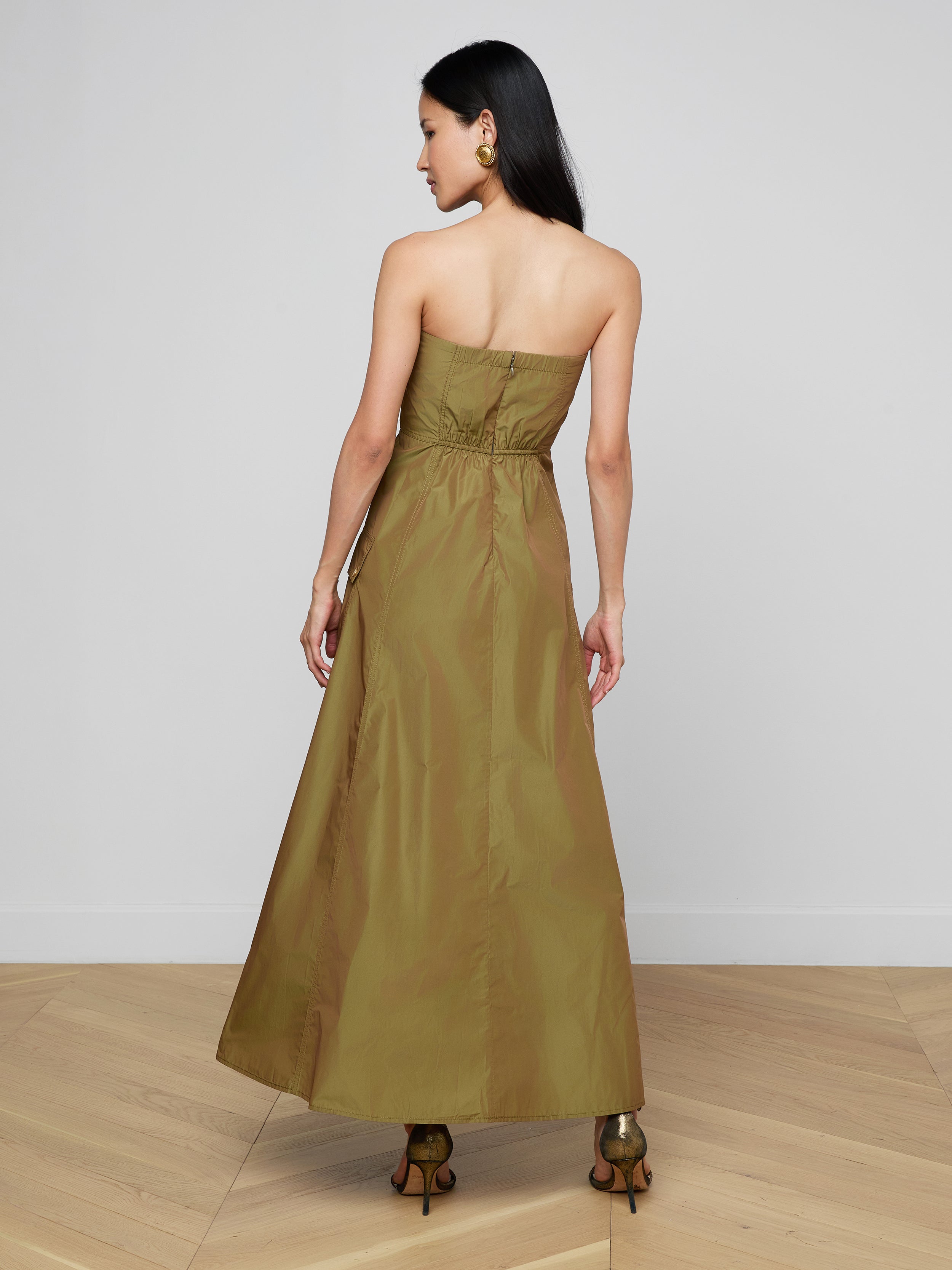 A woman stands facing away, wearing the Nessa Strapless Gown—an olive-green, floor-length dress featuring a fitted bodice and a flared skirt. She has long dark hair, visible earrings, and brown heels, set against a plain white background.