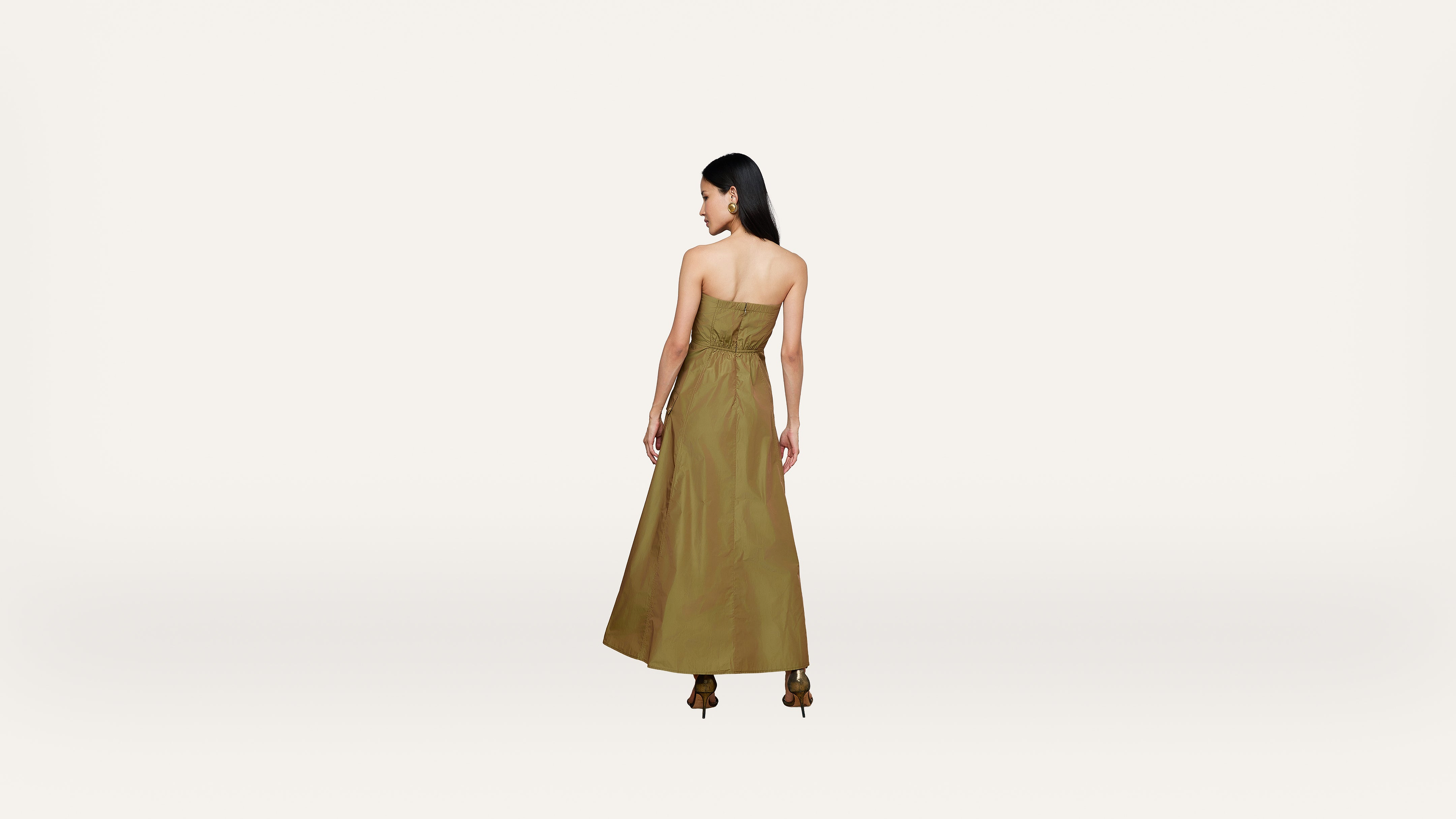 A woman with long dark hair wears a strapless, olive green maxi dress and heels, standing with her back to the camera against a plain white background.