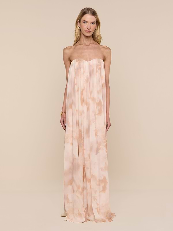 A woman stands against a plain white background, wearing the Raine Strapless Gown in a light peach and cream tie-dye design. The floor-length dress features a built-in corset, and she faces forward with a neutral expression, her arms at her sides.