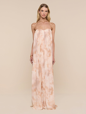 A woman with straight blonde hair and minimal jewelry stands against a beige background in the Raine Strapless Gown, a floor-length peach and cream tie-dye dress featuring a built-in corset, exuding relaxed elegance.