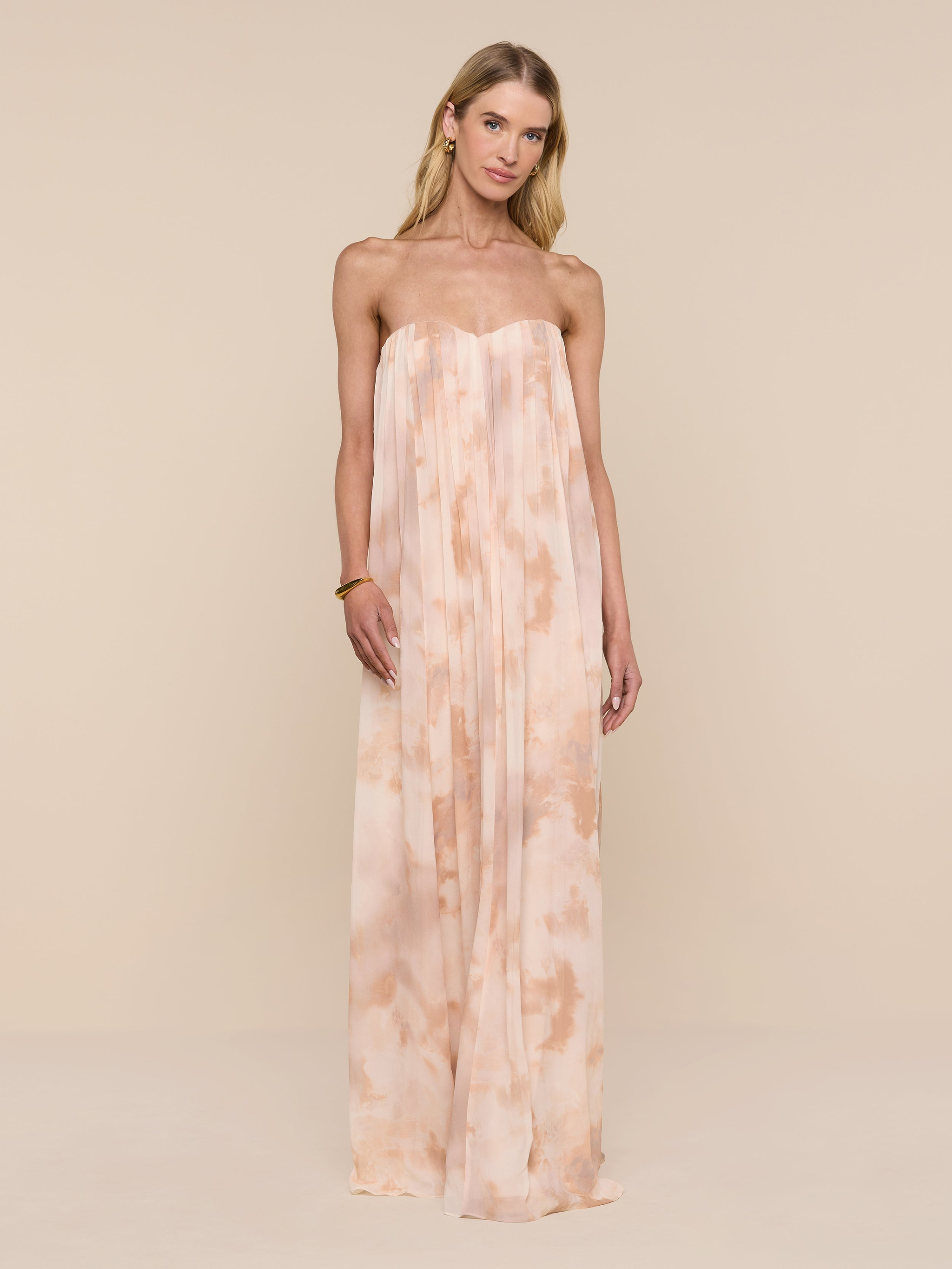 A woman stands against a plain white background wearing the Raine Strapless Gown, featuring flowing peach and cream tie-dye fabric and a built-in corset.